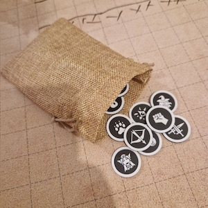 Può includere: Un sacco di iuta beige aperto, che versa gettoni di gioco in bianco e nero. I gettoni presentano vari simboli, tra cui un'impronta di zampa, arco e freccia e un pugno. I gettoni sono sparsi su una superficie beige.