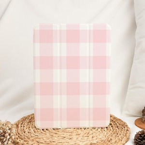 May include: A tablet case with a pink and white gingham pattern. The case is rectangular and has a folding cover. It is placed on a round, woven placemat. The background is white fabric.