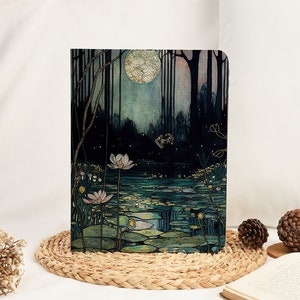Puede incluir: Un cuaderno negro y verde con una escena de bosque iluminado por la luna. La portada presenta una luna llena, árboles y un estanque con nenúfares y flores. La escena está pintada en un estilo acuarela con detalles dorados.