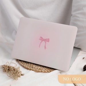 May include: A light pink laptop with a watercolor-style pink bow decal. The laptop is resting on a woven mat, with a white blanket in the background. The bow is centered on the laptop's lid.