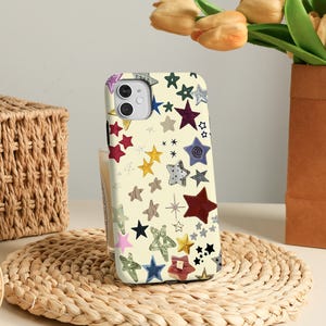 Puede incluir: Una funda de teléfono con un fondo color crema y un patrón de estrellas coloridas de varios tamaños y diseños. Las estrellas son en tonos de rojo, azul, verde, amarillo y gris. La funda está sobre una estera tejida.