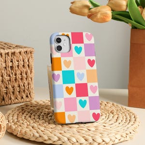 Könnte beinhalten: Eine bunte Handyhülle mit einem Schachbrettmuster aus Quadraten und Herzen in Orange-, Rosa-, Lila-, Blau- und Weißtönen. Die Hülle liegt auf einer gewebten Matte, mit einem Weidenkorb und einer Vase mit Blumen im Hintergrund.