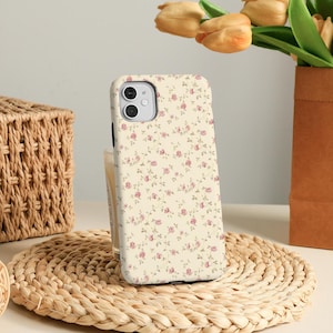 May include: A phone case with a floral pattern of small pink roses and green leaves on a cream background. The case is on a woven mat, with a wicker basket and a vase of tulips in the background.