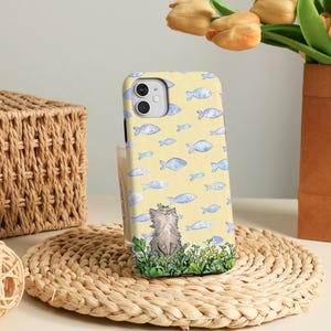 May include: A yellow phone case with a pattern of blue fish. The case has a watercolour illustration of a grey cat wearing a leaf crown, sitting in green grass. The case is on a woven mat.