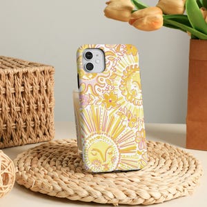 May include: A yellow and white floral patterned phone case with a sun design. The case is on a woven coaster with a basket and a decorative ball in the background. A vase of yellow flowers is also visible.