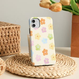May include: A phone case with a cream-colored background and a pattern of colorful flowers in orange, pink, yellow, green, and blue. The case is on a woven straw mat, with a woven basket and a vase of flowers in the background.