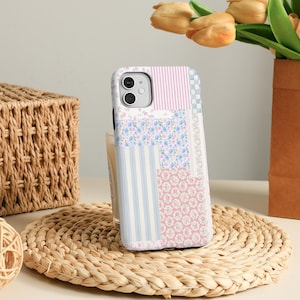 May include: A patterned phone case featuring a patchwork design with pink stripes, blue floral, and white and blue stripes. The case is displayed on a woven mat, with a basket and flowers in the background.