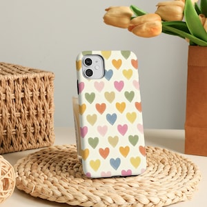Puede incluir: Funda de teléfono con un patrón de corazones multicolores sobre un fondo crema. La funda está colocada sobre un salvamanteles tejido, con una cesta y flores en el fondo. Los corazones son en tonos de amarillo, naranja, rosa, verde y azul.