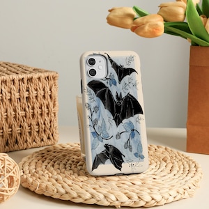 Op de afbeelding: Een crèmekleurige telefoonhoes met een ontwerp van zwarte vleermuizen en blauwe en witte botanische accenten. De hoes heeft een glanzende afwerking en een camera-uitsparing.