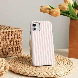 Könnte beinhalten: Eine Smartphone-Hülle mit vertikalem rosa-weißem Streifenmuster. Die Hülle liegt auf einem gewebten Platzset, im Hintergrund ein Weidenkorb und ein dekorativer Ball. Ein Strauß gelber Tulpen steht in einer braunen Papiervase.
