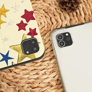 May include: A clear phone case next to a phone case with a star design. The star design includes gold, red, blue, and silver stars on a cream background. The phone cases are on a woven, light brown surface.