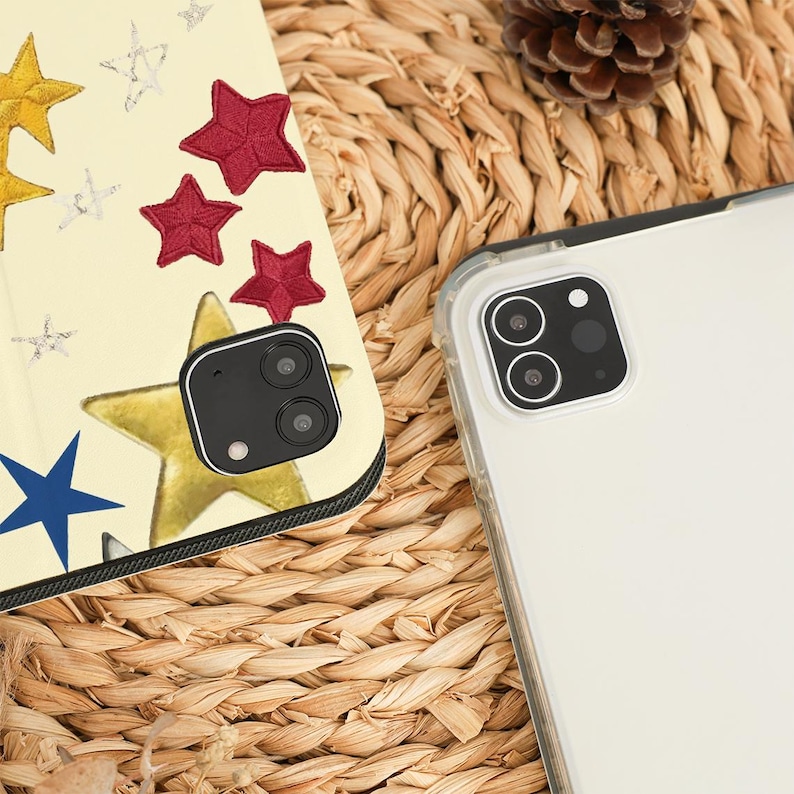 May include: A clear phone case next to a phone case with a star design. The star design includes gold, red, blue, and silver stars on a cream background. The phone cases are on a woven, light brown surface.