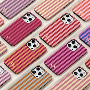 Op de afbeelding: Een collectie smartphonehoesjes met verticale strepen in diverse kleurcombinaties, waaronder roze, rood, oranje, geel en blauw. Elk hoesje heeft een uitsparing voor de camera en goudkleurige initialen op de onderkant van de achterzijde.
