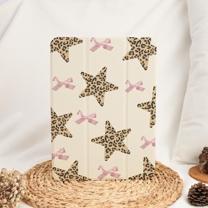 Può includere: Custodia per tablet color crema con un motivo ripetuto di stelle con stampa leopardata e piccoli fiocchi rosa. La custodia è in piedi su un tappetino intrecciato, mostrando il suo design. La custodia ha una chiusura magnetica.