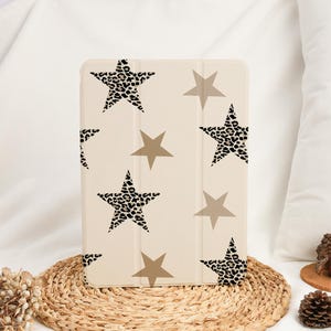 Puede incluir: Una funda beige para tableta con un estampado de estrellas marrones y de leopardo. La funda es rectangular con una tapa plegable. Las estrellas son de diferentes tamaños y están espaciadas uniformemente.