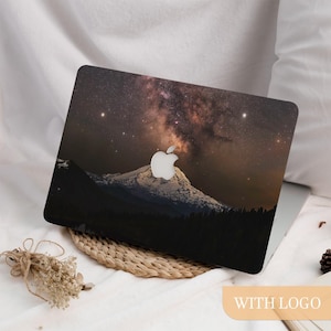 May include: A laptop skin featuring a night sky with a visible Milky Way galaxy, stars, and a snow-capped mountain peak. The design includes a central cutout for the Apple logo, showcasing a dark, celestial landscape with deep brown and black tones.