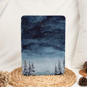 以下が含まれることがあります： 水彩画の冬の風景が描かれたタブレットケース。デザインは、白い星と雪に覆われた常緑樹のある濃い青色の夜空を示しています。長方形のケースは、耐久性のある素材で作られているようです。