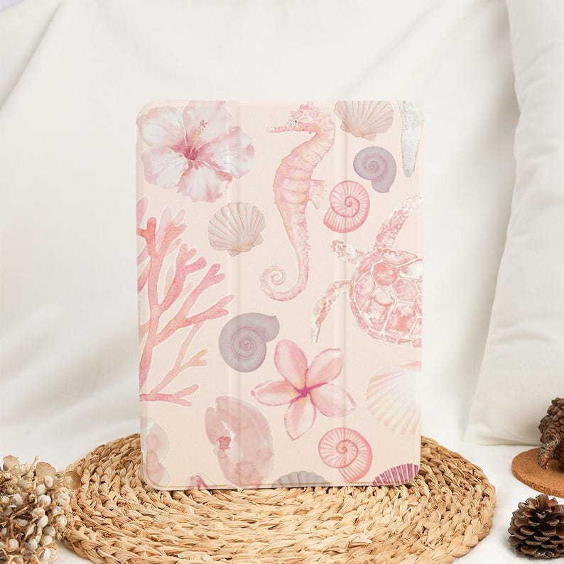 Pu&ograve; includere: Una custodia rettangolare per tablet con sfondo rosa chiaro e un motivo ad acquerello ripetuto raffigurante cavallucci marini, tartarughe marine, varie conchiglie, rami di corallo e fiori di ibisco nei toni del rosa e del bianco.