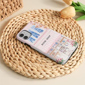 May include: A phone case featuring a pink and white illustration of a bookstore with the text "BOOK SHOP". The case has a detailed design of windows, bookshelves, and floral accents. The phone case is resting on a woven mat.