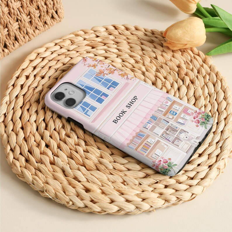 May include: A phone case featuring a pink and white illustration of a bookstore with the text "BOOK SHOP". The case has a detailed design of windows, bookshelves, and floral accents. The phone case is resting on a woven mat.