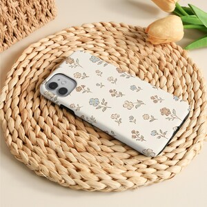 K&ouml;nnte beinhalten: Eine wei&szlig;e Smartphone-H&uuml;lle mit einem wiederkehrenden Muster aus kleinen, zarten Blumen in den Farben Hellblau, sanftem Braun und gedecktem Beige. Die H&uuml;lle hat einen rechteckigen Ausschnitt f&uuml;r eine Doppelkamera und einen schwarzen Rand.