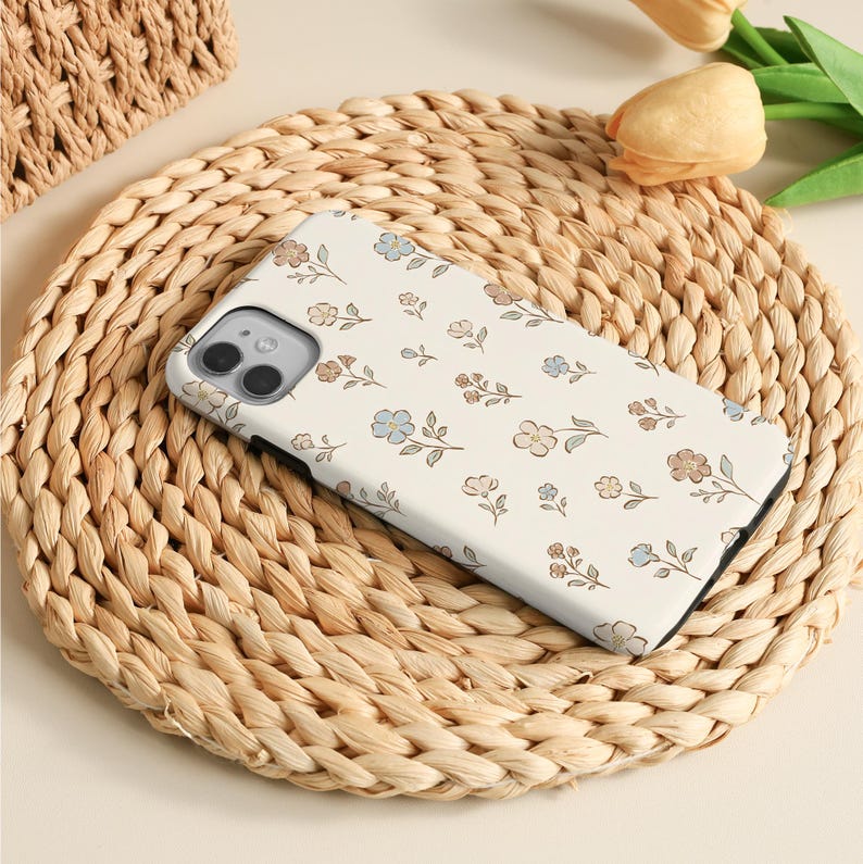 K&ouml;nnte beinhalten: Eine wei&szlig;e Smartphone-H&uuml;lle mit einem wiederkehrenden Muster aus kleinen, zarten Blumen in den Farben Hellblau, sanftem Braun und gedecktem Beige. Die H&uuml;lle hat einen rechteckigen Ausschnitt f&uuml;r eine Doppelkamera und einen schwarzen Rand.