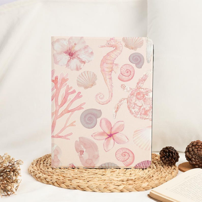 Pu&ograve; includere: Un diario con una copertina rosa chiaro con illustrazioni ad acquerello di un cavalluccio marino, conchiglie, coralli e fiori. Il design ha un'estetica morbida e femminile. Il diario &egrave; esposto su un sottobicchiere intrecciato.