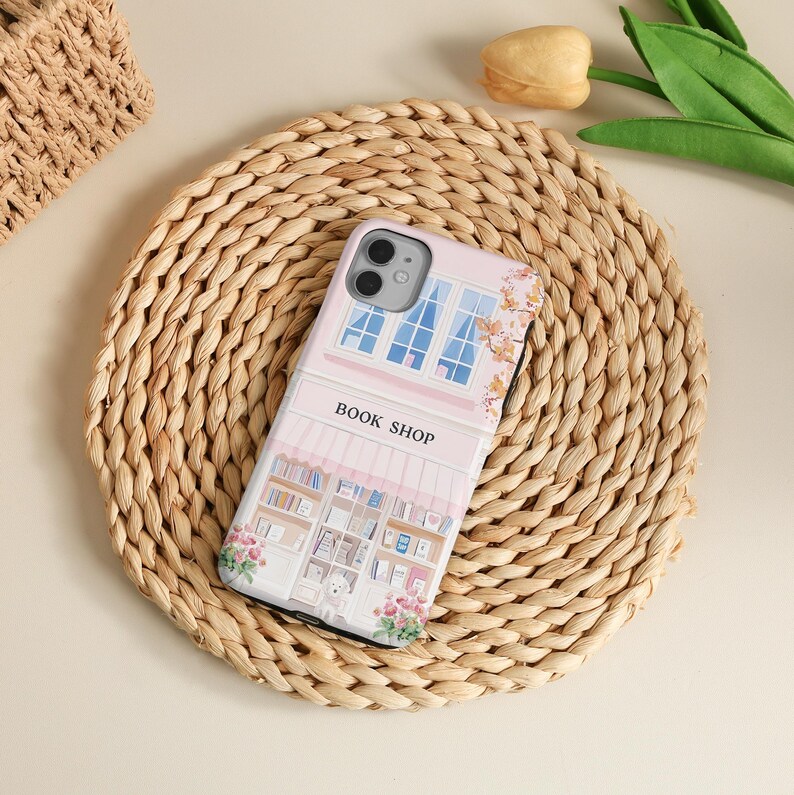 May include: A pink phone case featuring a book shop design. The case has a detailed illustration of a bookstore facade with windows, books, and floral accents. The words "BOOK SHOP" are printed on the case.