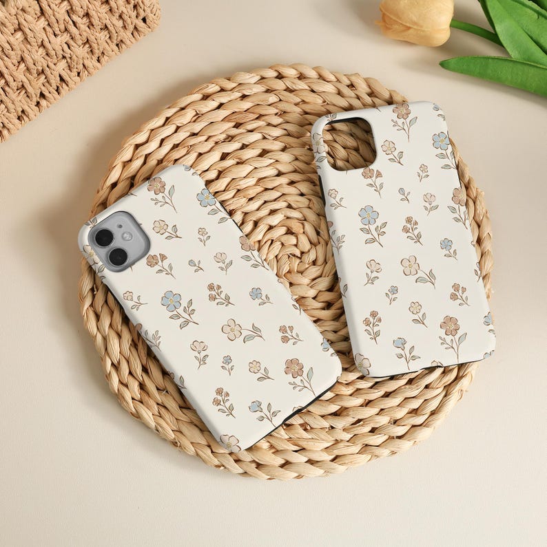 K&ouml;nnte beinhalten: Zwei cremefarbene Smartphone-H&uuml;llen mit einem wiederkehrenden Muster aus kleinen, zarten Blumen in gedeckten Blau-, sanften Braun- und hellen Beiget&ouml;nen. Jede H&uuml;lle hat einen rechteckigen Ausschnitt f&uuml;r die Kamera und eine glatte, matte Oberfl&auml;che.