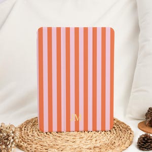 Peut inclure: Une coque de tablette rectangulaire présentant un motif à rayures verticales alternant des tons orange vif et rose clair. Le bas du centre de la coque affiche les lettres capitales dorées JM dans une police serif, centrées sur les rayures.