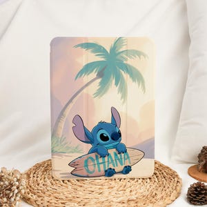 May include: A rectangular tablet case featuring a graphic illustration of a blue alien character holding a surfboard with the text OHANA printed on it. The background depicts a beach scene with a palm tree under a soft, warm-toned sky.