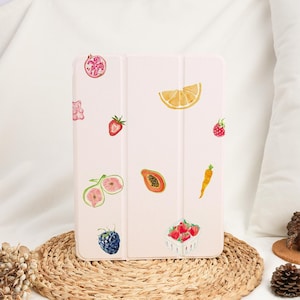 May include: A light pink tablet case adorned with watercolor illustrations of various fruits, including strawberries, a lemon slice, and a papaya. The case is resting on a woven mat, with a white background.