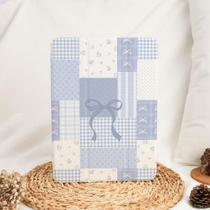 Puede incluir: Una funda para tableta de retazos azul claro y blanco con un diseño de lazo. La funda presenta varios patrones, incluyendo cuadros, lunares e estampados florales. La funda se muestra sobre una estera tejida.