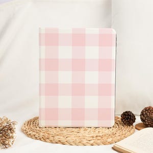 Pu&ograve; includere: Un quaderno con motivo a quadri rosa e bianco. Il quaderno &egrave; rettangolare e ha una copertina rigida. &Egrave; appoggiato su un sottobicchiere intrecciato. Lo sfondo &egrave; un morbido tessuto bianco.