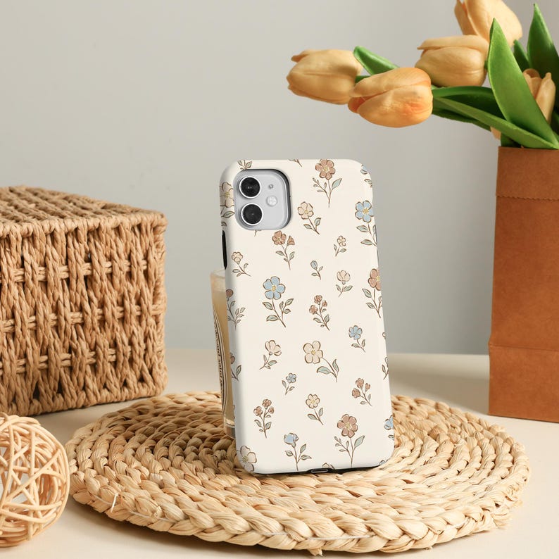 K&ouml;nnte beinhalten: Eine cremefarbene Smartphone-H&uuml;lle mit einem wiederkehrenden Muster aus kleinen, zarten Blumen in Hellblau, sanftem Braun und ged&auml;mpftem Orange, wobei d&uuml;nne gr&uuml;ne St&auml;ngel und Bl&auml;tter &uuml;ber die gesamte R&uuml;ckseite der H&uuml;lle verteilt sind.