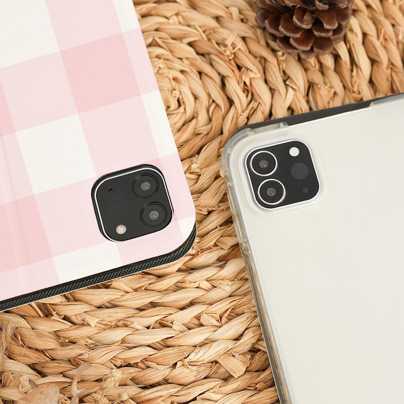 Pu&ograve; includere: Primo piano di due custodie per telefono. Una custodia ha un motivo a quadri rosa e bianco, l'altra &egrave; trasparente. Entrambe le custodie hanno ritagli per la fotocamera. Le custodie sono su una superficie tessuta con una pigna.