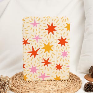 Peut inclure: Une coque de tablette avec un motif d'éclat d'étoiles colorées. Le fond crème est parsemé de tirets jaunes et d'étoiles vives rouges, roses et jaunes. La coque rectangulaire semble être faite d'un matériau durable, avec un design joyeux.