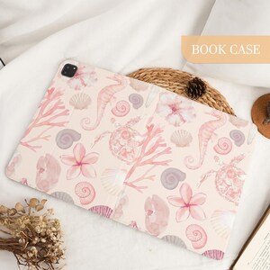 Pu&ograve; includere: Una custodia rettangolare per tablet con sfondo rosa chiaro e un motivo ad acquerello ripetuto di cavallucci marini, tartarughe marine, varie conchiglie e fiori tropicali. La custodia include un ritaglio per fotocamera e il testo BOOK CASE.