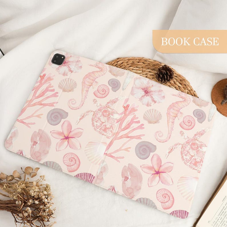 Pu&ograve; includere: Una custodia rettangolare per tablet con sfondo rosa chiaro e un motivo ad acquerello ripetuto di cavallucci marini, tartarughe marine, varie conchiglie e fiori tropicali. La custodia include un ritaglio per fotocamera e il testo BOOK CASE.