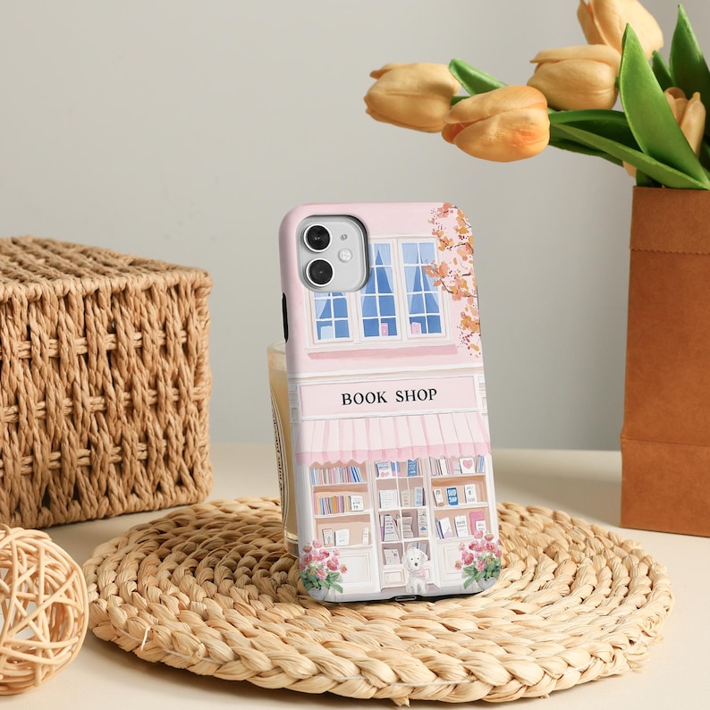 May include: A pink phone case featuring a whimsical illustration of a bookstore facade. The design includes a window, awning, and the words "BOOK SHOP". The case is decorated with floral accents and a small dog.