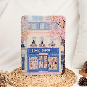 Może przedstawiać: Etui na tablet z szczegółową ilustracją niebieskiego sklepu "BOOK SHOP" i różowego kwitnącego drzewa. Okna sklepu prezentują rzędy książek, a budynek ma jasnoniebieską fasadę. Etui jest umieszczone na tkanej macie.