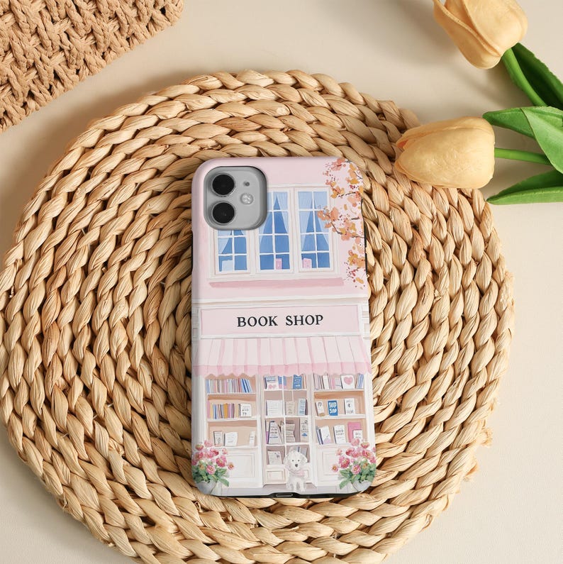May include: A pink phone case featuring a whimsical illustration of a bookstore. The design includes a storefront with a striped awning, bookshelves, and the text "BOOK SHOP". The case is decorated with floral accents and a small dog.