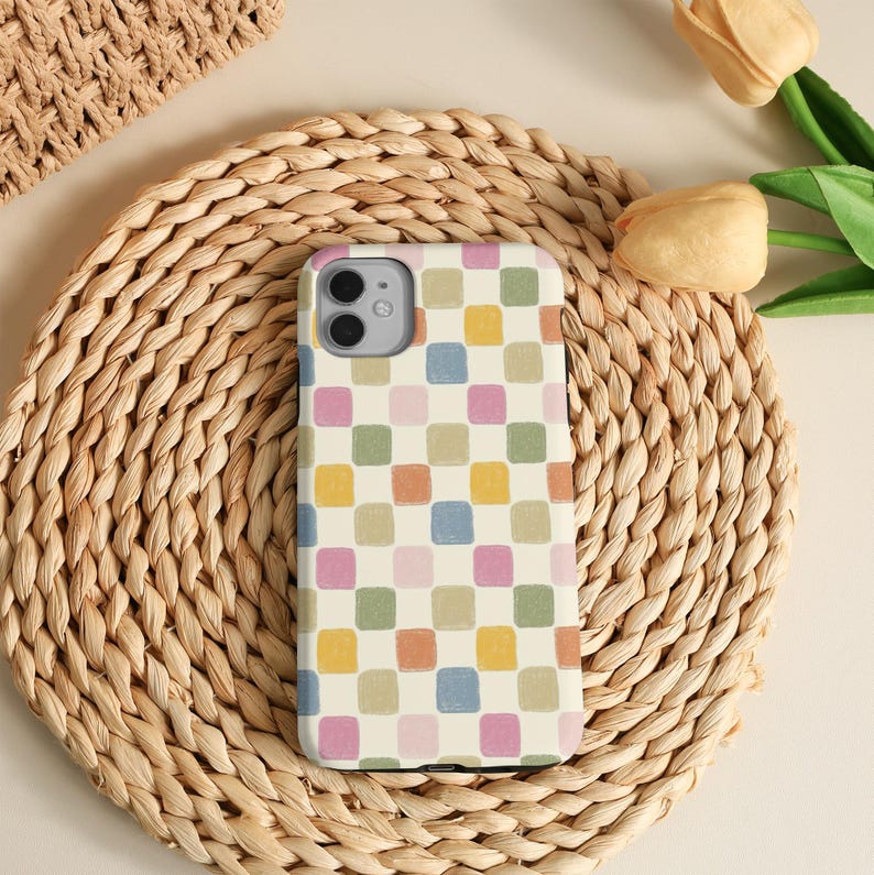 K&ouml;nnte beinhalten: Eine Handyh&uuml;lle mit einem farbenfrohen Schachbrettmuster in Pastellt&ouml;nen von Rosa, Gr&uuml;n, Orange, Gelb und Blau. Die H&uuml;lle liegt auf einem gewebten, kreisf&ouml;rmigen Platzdeckchen, mit einem Teil eines gewebten Korbes sichtbar.
