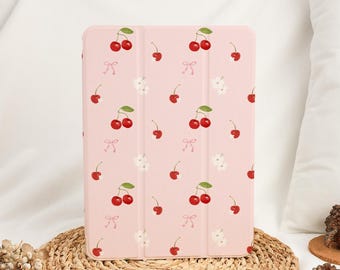 May include: A pink tablet case with a repeating pattern of red cherries, white flowers, and small pink bows. The case is rectangular and appears to be made of a smooth material. The case is sitting on a woven, circular mat.