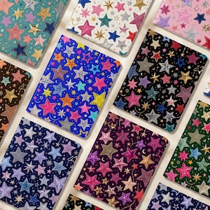 May include: A collection of rectangular notebooks featuring various background colors including teal, white, pink, blue, black, purple, and green, all decorated with a repeating pattern of multi-colored, glittery, and metallic stars.