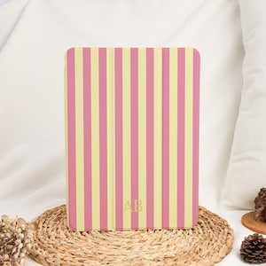 Pode incluir: Uma capa retangular para tablet com um padrão de riscas verticais em tons alternados de amarelo pálido e rosa seco. A parte central inferior da capa apresenta as letras maiúsculas douradas AB numa fonte serifada, centradas nas riscas.