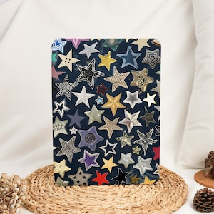 Puede incluir: Un panel decorativo rectangular con un fondo azul oscuro cubierto con varias formas de estrellas en diferentes colores, texturas y tamaños. Algunas estrellas tienen un brillo metálico, mientras que otras son de papel o cerámica. El texto "STAR GIRL" es visible.