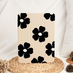 Puede incluir: Una funda beige para tableta con un patrón repetido de grandes flores negras con centros blancos. La funda es rectangular y parece estar hecha de un material liso. El diseño es simple y moderno.