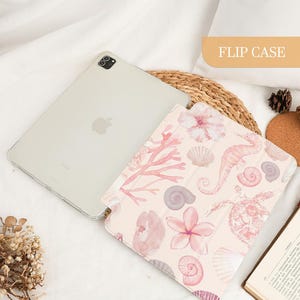 Pu&ograve; includere: Una custodia a libro per tablet con sfondo rosa chiaro e un motivo di illustrazioni ad acquerello raffiguranti cavallucci marini, tartarughe marine, varie conchiglie, coralli e fiori di ibisco. La custodia &egrave; aperta accanto a un tablet argentato.
