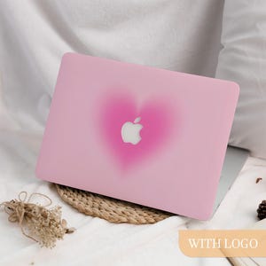 Peut inclure: Une coque d'ordinateur portable rose avec un motif de cœur flou rose foncé. Le logo Apple est visible au centre du cœur. La coque est posée sur un tapis tressé, avec des fleurs séchées et le texte "WITH LOGO".